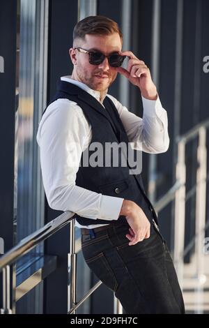Man in elegant clothes and sunglasses is standing indoors and posing for the camera Stock Photo