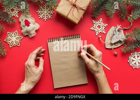 Woman writing Christmas whishes in craft sketchbook, notebook on red background. View from above. Flat lay. Space for text. Stock Photo