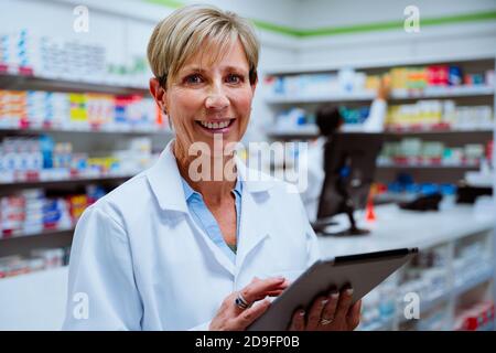 Smiling senior pharmacist scrolling through digital tablet standing in pharmacy drugstore  Stock Photo