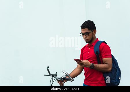 Latin young man with a backpack and glasses looking at his phone and standing near the bicycle Stock Photo
