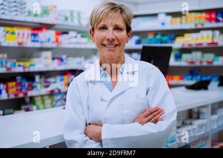 Portrait of smiling caucasian pharmacist with crossed arms leaning against medication counter in pharmacy  Stock Photo