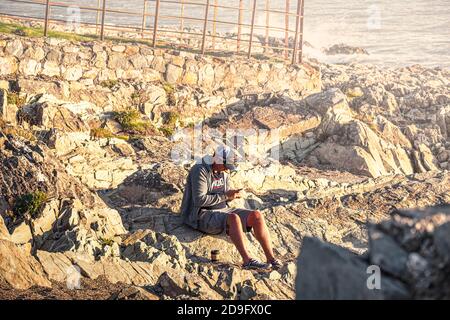 Everyday life. Man with mobile phone sitting on rocks near the sea. Stock Photo
