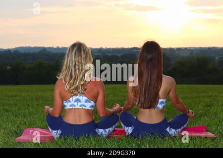Two girls sitting in a yoga pose, one blond and one brunette on a mat outdoors with the sun setting in front of them Stock Photo