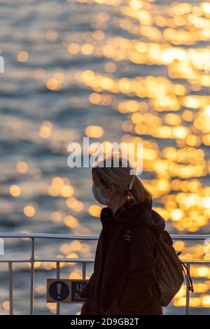 a woman sitting next to some railings next to the sea at sunset looking out over the golden coloured water daydreaming. Stock Photo