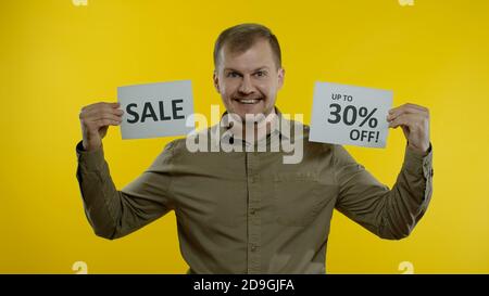 Great discounts. Attractive joyful blonde man showing Sale word and showing Up To 30 percent Off purchase inscription. Shopping with low prices, Black Friday concept. Yellow background Stock Photo