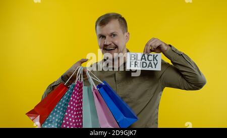 Happy man with shopping bags showing Black Friday inscription note, smiling, rejoicing good discounts, low prices for online shopping sales. Studio shot on yellow background Stock Photo