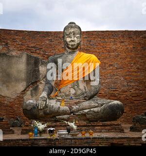 A seated buddha statue wearing a saffron sash in a brick courtyard of a thai buddhist temple. In front of the statue are a variety of offerings Stock Photo