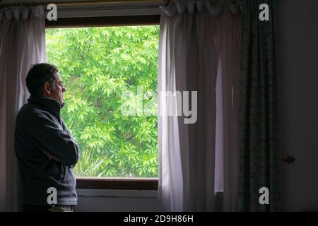 Pensive senior adult man looking out the window , with a background of green leaves Stock Photo
