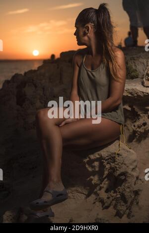 Pretty woman in a T-shirt and swimsuit, looking at a beautiful sunset by the sea, sitting on a rock. Long shot. Formentera island, Spain. Stock Photo