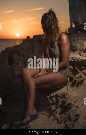 Pretty woman in a T-shirt and swimsuit, looking at a beautiful sunset by the sea, sitting on a rock. Long shot. Formentera island, Spain. Stock Photo