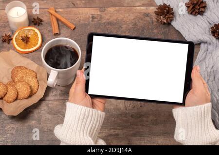 Mockup tablet pc. Woman's hands holding black tablet pc with blank white screen while drinking coffee on wooden table in cafe. winter Christmas time Stock Photo