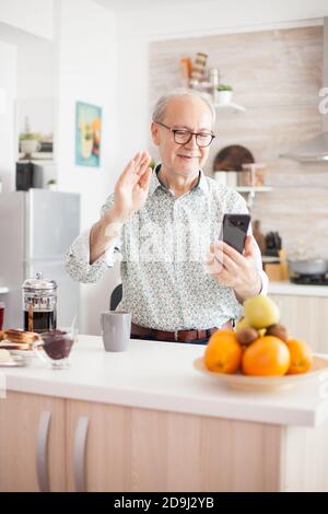Senior man having a conversation, using his modern phone. Elderly aged man waving during a video conference with family in kitchen. Elderly person using internet online chat technology video webcam making a video call connection camera communication conference call Stock Photo
