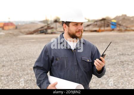 Portrait of foreman standing on construction site, talking by VHF walkie talkie and holding blueprints. Stock Photo