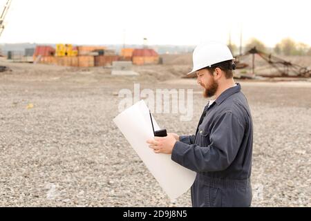 Back view of male building engineer standing on construction site, talking by VHF walkie talkie and holding blueprints. Stock Photo