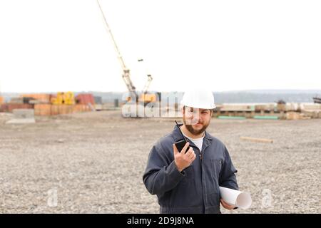 Foreman standing on construction site, talking by VHF walkie talkie and holding blueprints. Stock Photo
