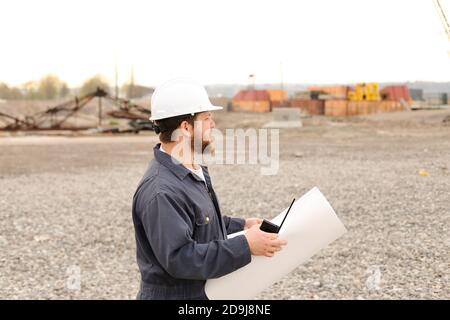 Male building engineer standing on construction site, talking by VHF walkie talkie and holding blueprints. Stock Photo