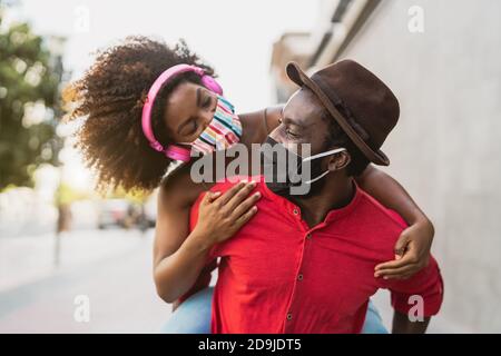 African couple piggyback wearing face mask - Happy Afro people having fun outdoor - Health care and youth relationship concept Stock Photo