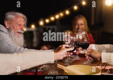 Happy multiracial seniors toasting with red wine glasses and celebrating holidays on house patio dinner - Elderly lifestyle people concept