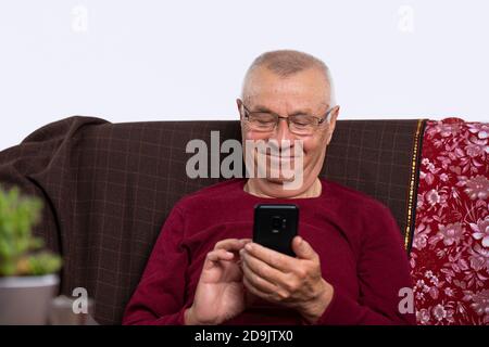 Happy elderly man in glasses holding smartphone while relaxing on the sofa at home. Modern technology concept. Stock Photo
