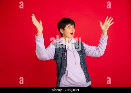 Shocked, astonished. Portrait of senior woman in stylish outfit, attire isolated on red studio background. Tech and joyful elderly lifestyle concept. Trendy colors, forever youth. Copyspace for your ad. Stock Photo