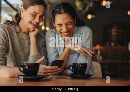 Two female friends sitting in a coffee shop and smiling. Two attractive women using a cellphone while relaxing together at a cafe. Stock Photo