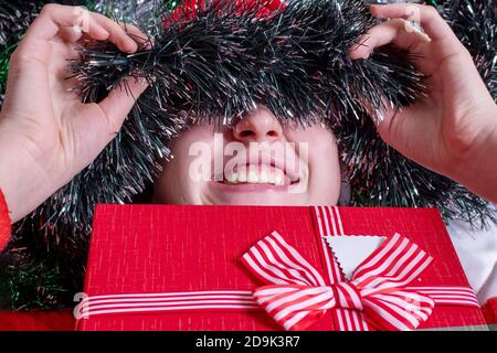 The joy of Christmas gifts. Close-up of a happy, smiling girl hiding behind green tinsel and a Christmas present. Happy face of a child close-up. Back Stock Photo