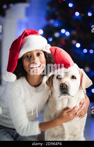 Smiling african american girl hugging labrador in santa hat on blurred background Stock Photo