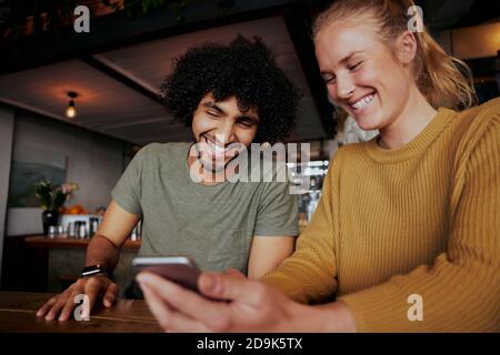 Cheerful male and female friends laughing looking at video in smartphone sitting in cafe Stock Photo