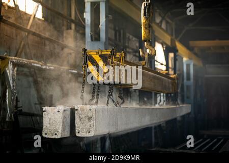 The crane moves a reinforced concrete product with holes. Reinforced concrete pillars fixed with metal hooks and chains on the background of the plant Stock Photo