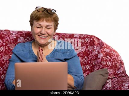 A nice-looking elderly woman, uses a laptop and smiles happily in video call. Photo of a house on a couch with an empty side space. Stock Photo