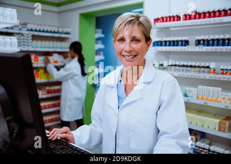 Caucasian senior pharmacist smiling while working on desktop computer searching for scripts standing in pharmacy  Stock Photo