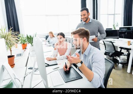 Businessman using graphics tablet and computer near multicultural colleagues in office Stock Photo