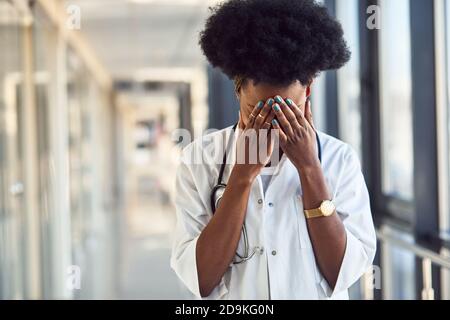 Sad and depressed young african american female doctor in white uniform standing in corridor Stock Photo