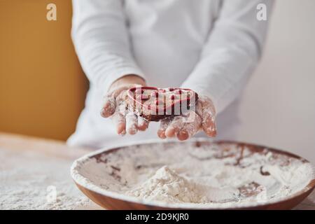 Close up view of little girl's hands that holding sweet cookie on the kitchen with plate of flour Stock Photo