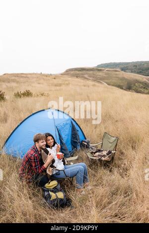 Smiling interracial couple with blanket and cup sitting near tent on grassy lawn Stock Photo