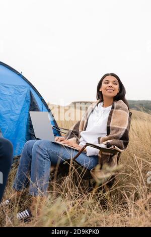 Smiling african american woman wrapped in blanket using laptop near tent on lawn Stock Photo