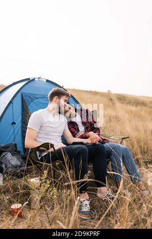 Smiling multiethnic couple holding hands while sitting near tent on lawn Stock Photo