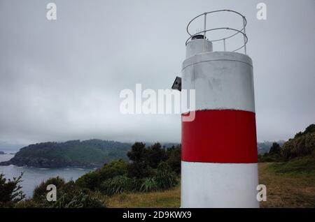 Small lighthouse with white and red stripes on a cape with view of overcast sky with fog over Pacific Coast. Bahia Mansa, Osorno, Chile Stock Photo