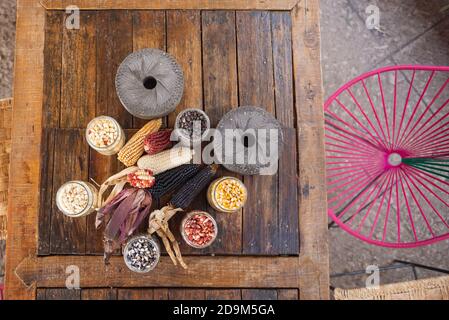 Colorful corn plant ears and kernels in glass jars with grinding stones on a wooden working table Stock Photo