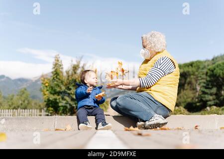 Grandmother wearing medical mask playing with leaves with her little grandson in a park Stock Photo