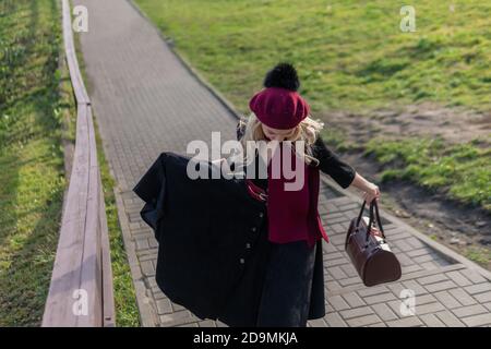 lady walks up the stairs and in burgundy palette and biret, an adult looking at the camera in black clothes, in the fall against a blue sky background Stock Photo