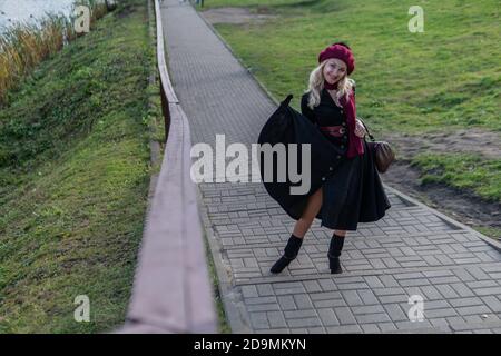 A cheerful lady walks up the stairs, both in burgundy palette and biret, an adult with gentle lips in black clothes, in the fall against the Stock Photo