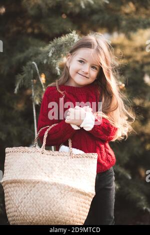 Smiling kid girl 3-4 year old wearing red knitted sweater holding straw basket over nature background outdoors close up. Childhood. Looking at camera. Stock Photo