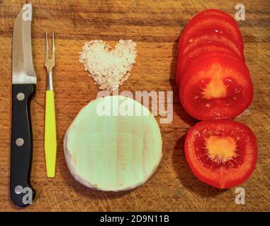 Sliced tomatoes with white mold cheese, heart-shaped stone salt and fork and knife on a cutting board. Stock Photo