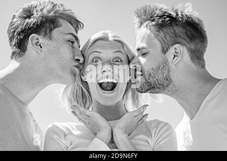Blonde happy girl getting kisses from two handsome boys. Charming woman standing between two friends. Best friends concept. Group of friends at clear Stock Photo