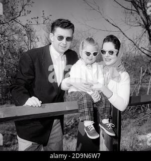 1950s FAMILY FATHER MOTHER DAUGHTER AT FENCE WEARING DARK SUNGLASSES LOOKING AT CAMERA - j2428 RGE001 HARS COUPLES HUSBAND 3 DAD FENCE MOM CLOTHING NOSTALGIC PAIR SUBURBAN SUNGLASSES RELATIONSHIP MOTHERS OLD TIME NOSTALGIA OLD FASHION 1 JUVENILE STYLE MYSTERY FAMILIES JOY LIFESTYLE PARENTING FEMALES VERTICAL MARRIED RELATION PORTRAITS RURAL SPOUSE HUSBANDS GROWNUP HEALTHINESS HOME LIFE COPY SPACE PEOPLE CHILDREN HALF-LENGTH DAUGHTERS PERSONS CARING CONFIDENCE FATHERS HUSBAND AND WIFE B&W MEN AND WOMEN PARENT AND CHILD EYE CONTACT HUSBANDS AND WIVES RELEASES PARENTS AND CHILDREN OPTICAL Stock Photo
