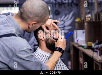 Young bearded man getting haircut by professional at modern barbershop. Beard and sideburns shaving and shaping with vintage straight razor. Movember. Stock Photo