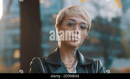 Portrait of fashionable caucasian confident blond woman with leather jacket and eyeglasses in the park. High quality photo