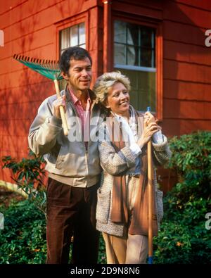 1980s MATURE COUPLE STANDING BY HOUSE MAN HOLDING RAKE ON SHOULDER WOMAN LEANING ON GARDEN TOOL WEARING AUTUMN CLOTHES SMILING - ks17849 PHT001 HARS BEAUTY TOOLS SUBURBAN COLOR EXPRESSION OLD TIME BUSY NOSTALGIA LEANING TOOL OLD FASHION 1 YARD FACIAL TEAMWORK PLEASED JOY LIFESTYLE CHORES FEMALES MARRIED SPOUSE HUSBANDS RAKE HEALTHINESS HOME LIFE COPY SPACE HALF-LENGTH LADIES PERSONS CARING MALES EXPRESSIONS MIDDLE-AGED PARTNER MIDDLE-AGED MAN HAPPINESS MIDDLE-AGED WOMAN WELLNESS CHEERFUL EXTERIOR PRIDE SMILES CONCEPTUAL JOYFUL STYLISH OWNERS TOGETHERNESS WIVES CAUCASIAN ETHNICITY OLD FASHIONED Stock Photo