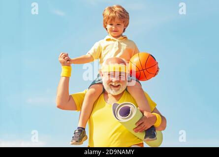 Preparing for morning workout in the park. Happy grandfather giving grandson piggyback ride on his shoulders and looking up. Stock Photo
